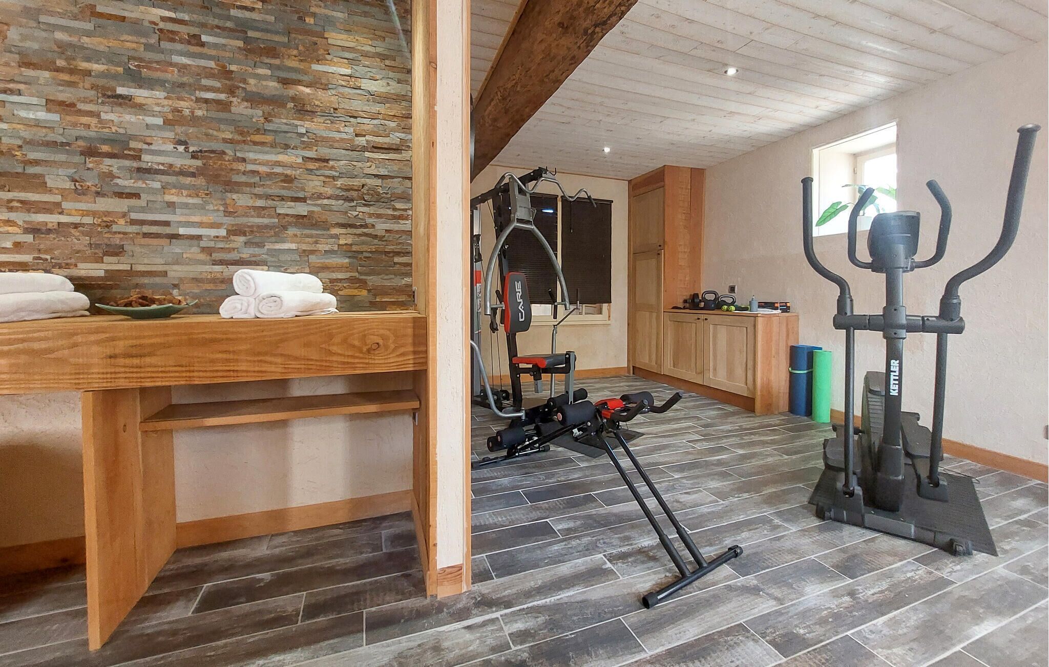 Quarto com parede de pedra, móveis de madeira, equipamentos de ginástica e uma janela.