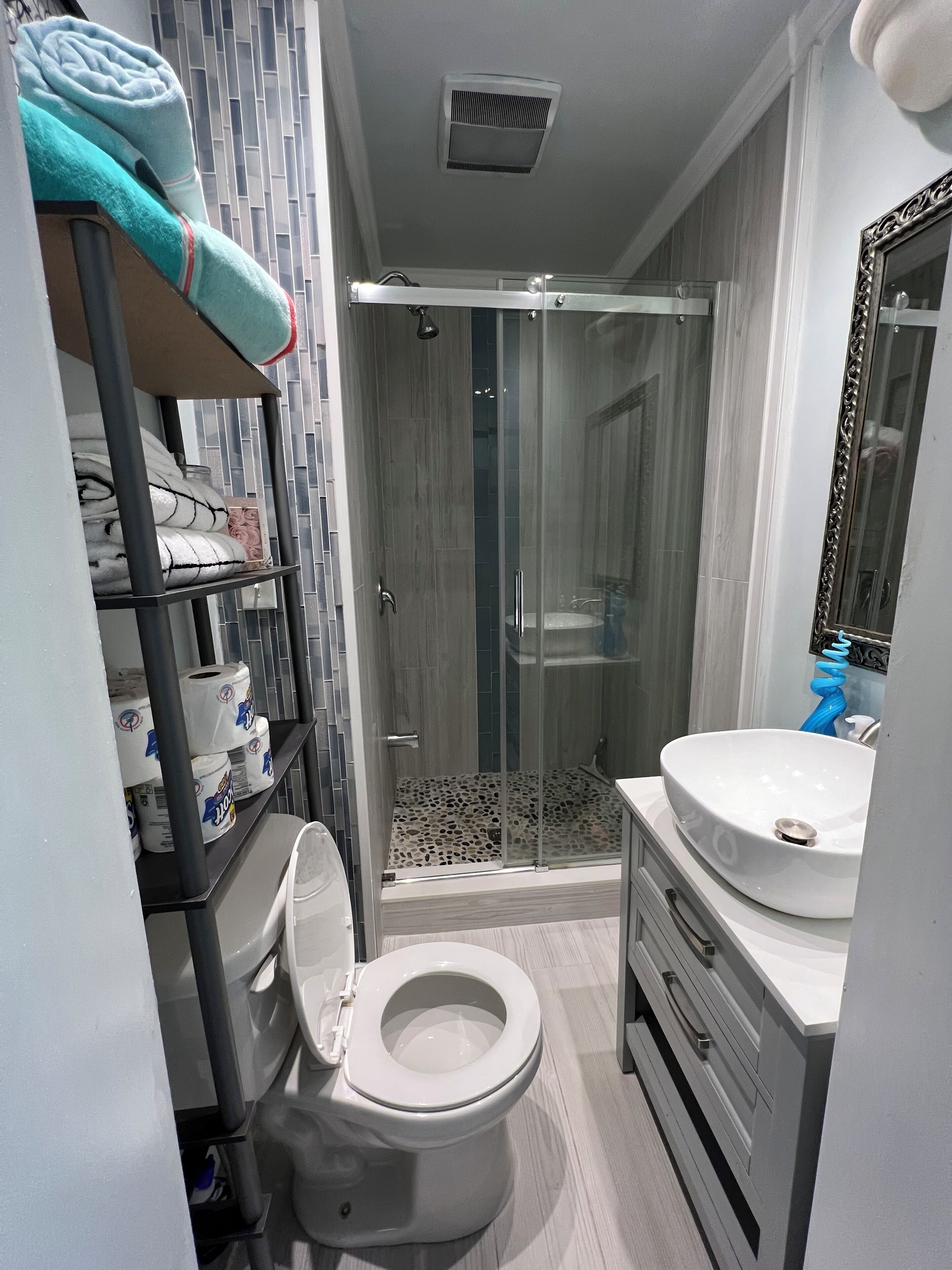 A bathroom with a toilet, a shower with a glass door, a sink with a mirror, and shelves with towels and toilet paper.