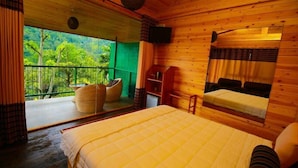 A wooden cabin room with a bed, a small table, a chair, a shelf, and a view of greenery outside. - Ella Taxi (Ella)