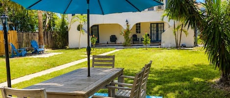 Un patio con una mesa y sillas de madera, una sombrilla azul y un edificio blanco al fondo.