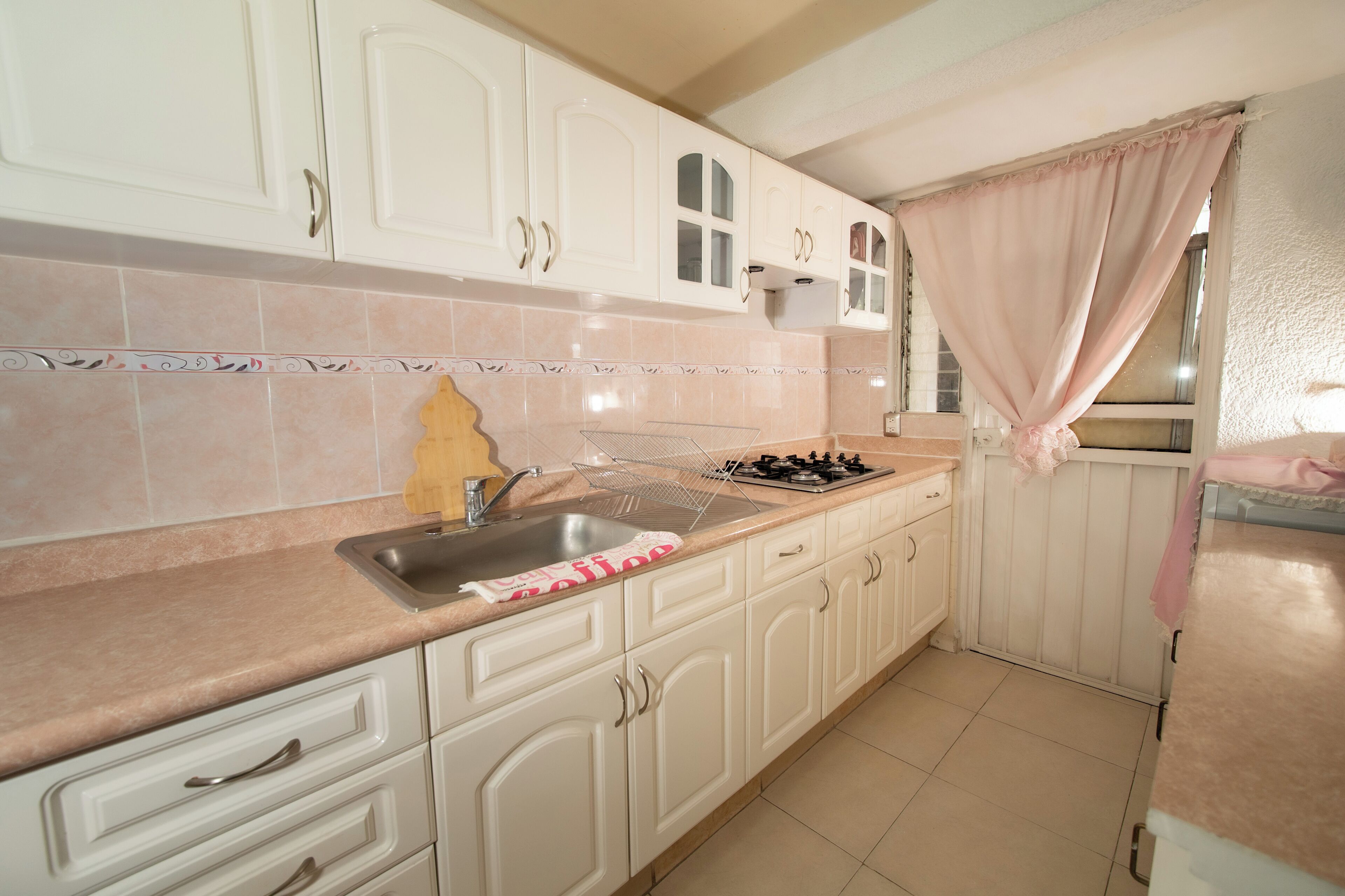 A kitchen with white cabinets, a stainless steel sink, a gas stove, and a window with a pink curtain.