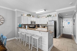 A kitchen with white cabinets, a stainless steel refrigerator, a microwave, and a coffee maker. There are three white bar stools and a clock on the wall. - Emerald Isle: Beachfront Condo, Pool, Balcony (Panama City Beach)