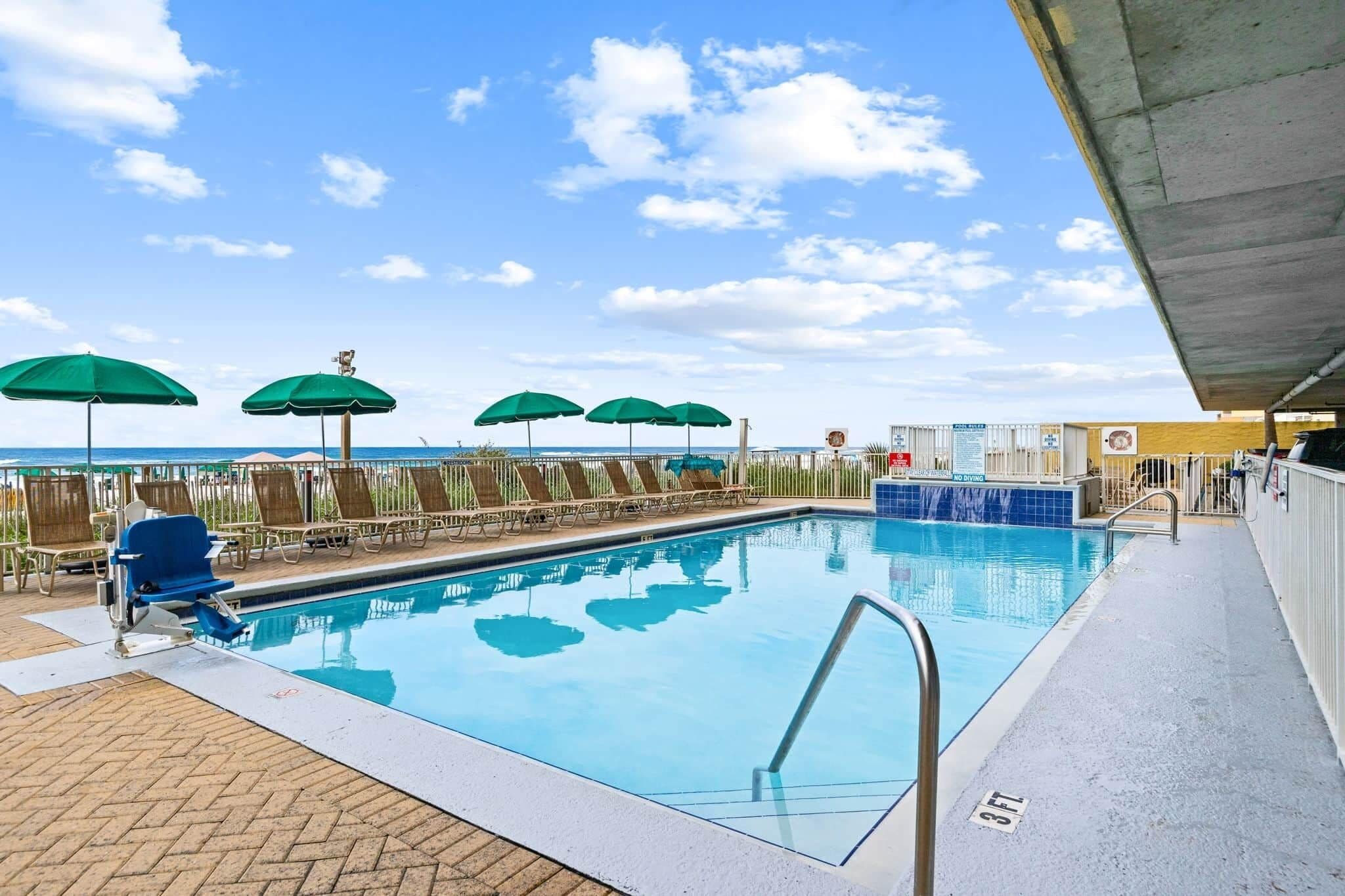 A swimming pool with lounge chairs and umbrellas, a diving board, and a view of the ocean.