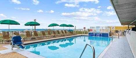 A swimming pool with lounge chairs and umbrellas, a diving board, and a view of the ocean.
