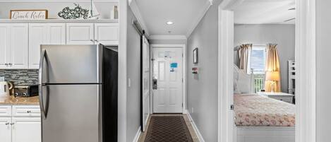Una cocina con un refrigerador de acero inoxidable, una puerta blanca con buzón y un pasillo que conduce a un dormitorio con cama y mesita de noche.
