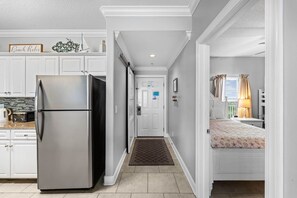 A kitchen with a stainless steel refrigerator, a white door with a mail slot, and a hallway leading to a bedroom with a bed and a nightstand. - Emerald Isle: Beachfront Condo, Pool, Balcony (Panama City Beach)