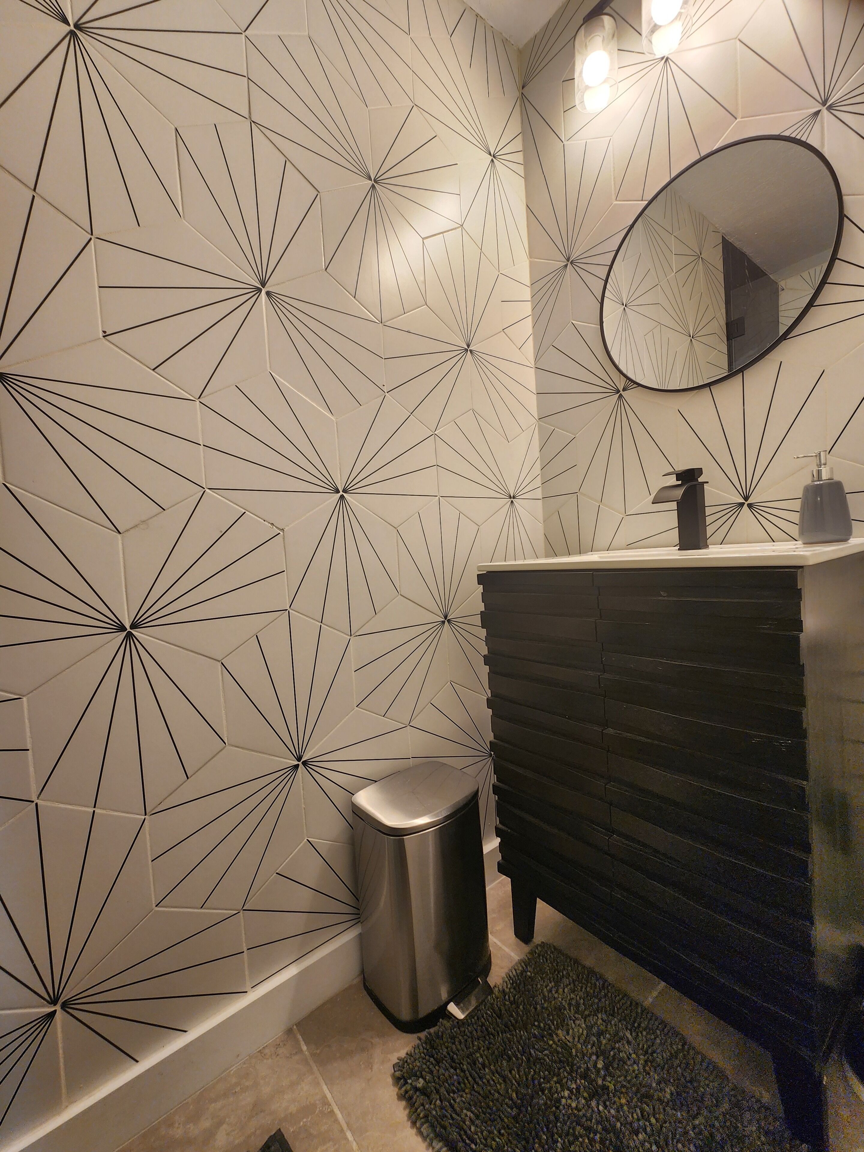 Un baño con pared de diseño geométrico, un espejo redondo, un lavamanos moderno y un cubo de basura.
