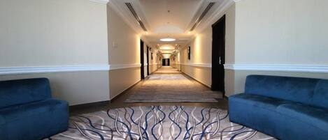 A hotel corridor with a carpeted floor, two sofas, and a door at the end.