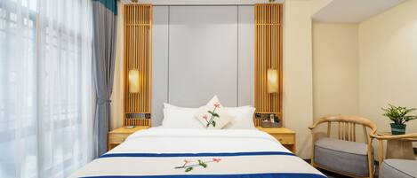 A hotel room with a bed, bedside tables, a chair, a small table, and a potted plant.