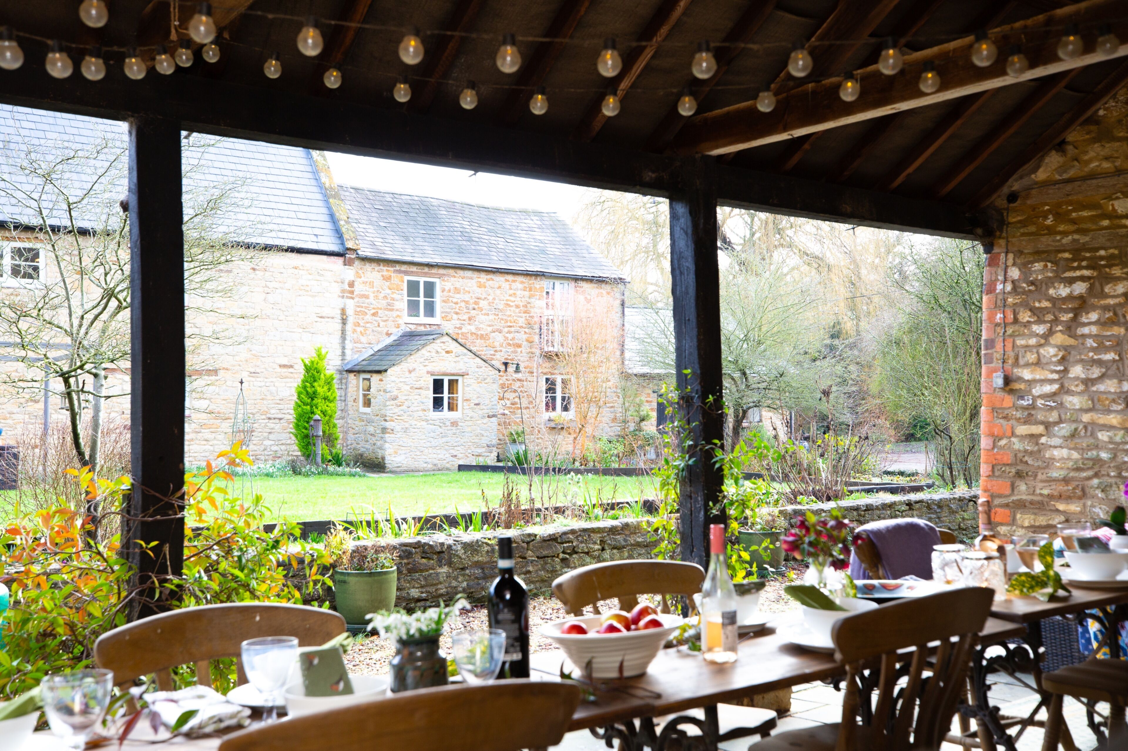 An outdoor dining area with stone walls, a view of a stone house, and a garden with autumn foliage.