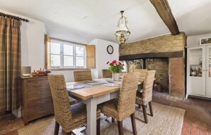 A dining area with a wooden table, wicker chairs, a hanging light fixture, a stone fireplace, and a window with curtains. - 17th Century Farmhouse - Near Silverstone Circuit (Towcester)