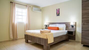 Een hotelkamer met een bed, nachtkastje, een wandairconditioner, een raam met gordijnen en een schilderij aan de muur.