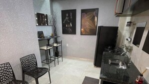 Een functionele keuken met een zwart aanrecht, een spoelbak en een kookplaat. Twee foto's in lijsten hangen aan de muur, een klein tafeltje met stoelen, en een decoratieve stoel met een geometrisch patroon.