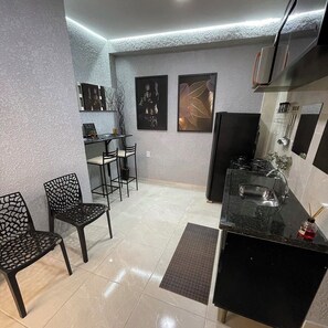 A compact kitchen with a black countertop, a sink, and a stove. There are two framed pictures on the wall, a small table with chairs, and a decorative chair with a geometric pattern. - Apartment in Copacabana Inhangá. In the best location in Rio de Janeiro, Copacabana. (Rio de Janeiro)