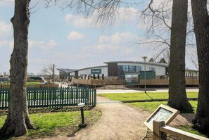 A building labeled 'The Venue' with a paved pathway leading to it, surrounded by trees and a green fence. - Billing Aquadrome Holiday Park - 2 Bedroom, Silver (Northampton)