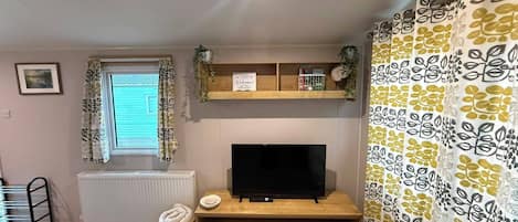 Una habitación con una televisión sobre un soporte de madera, un radiador abajo, una ventana con cortinas, una estantería con plantas y un letrero de bienvenida, además de una cortina estampada.