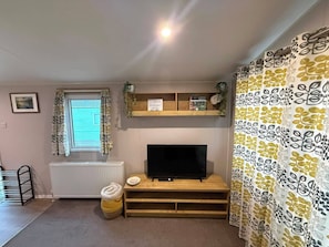 A room with a television on a wooden stand, a radiator below, a window with curtains, a shelf with plants and a welcome sign, and a patterned curtain. - Billing Aquadrome Holiday Park - 2 Bedroom, Silver (Northampton)