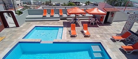 A rooftop pool area with two pools, lounge chairs, and umbrellas.