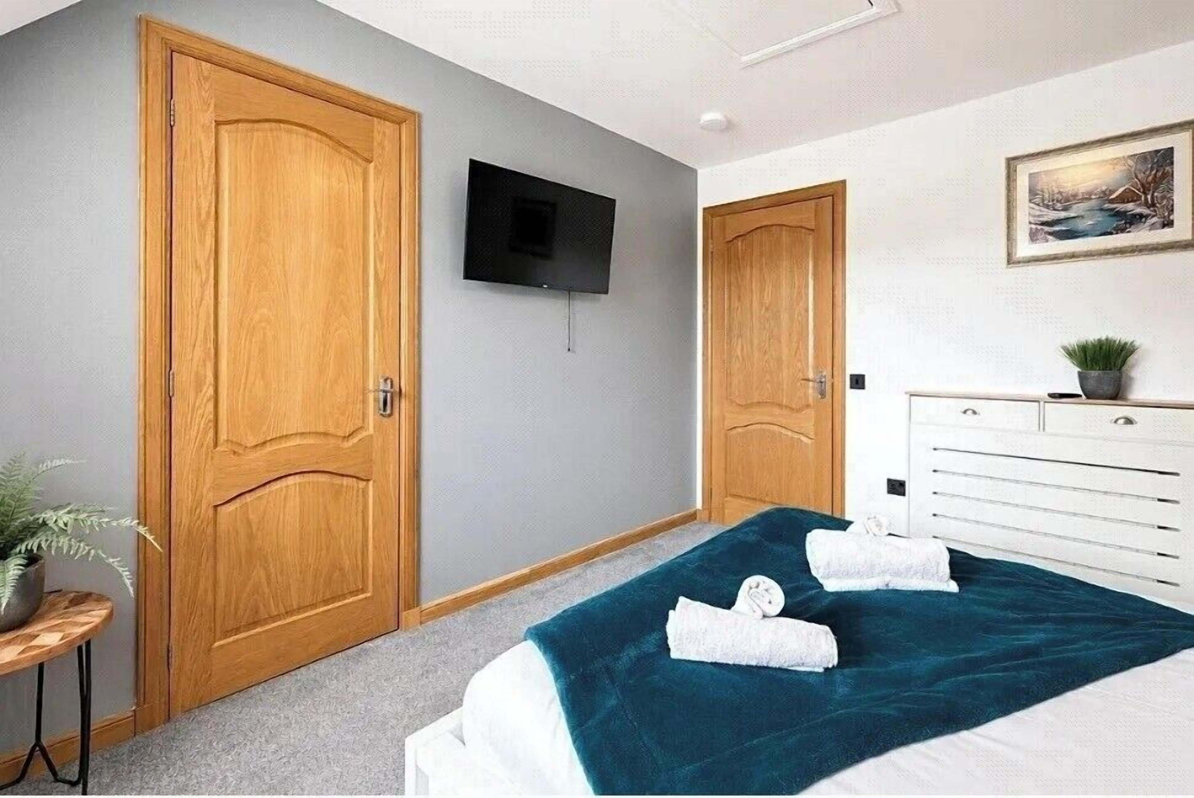 A hotel room with a bed, a television mounted on the wall, a wooden door, and a dresser with a plant on it.