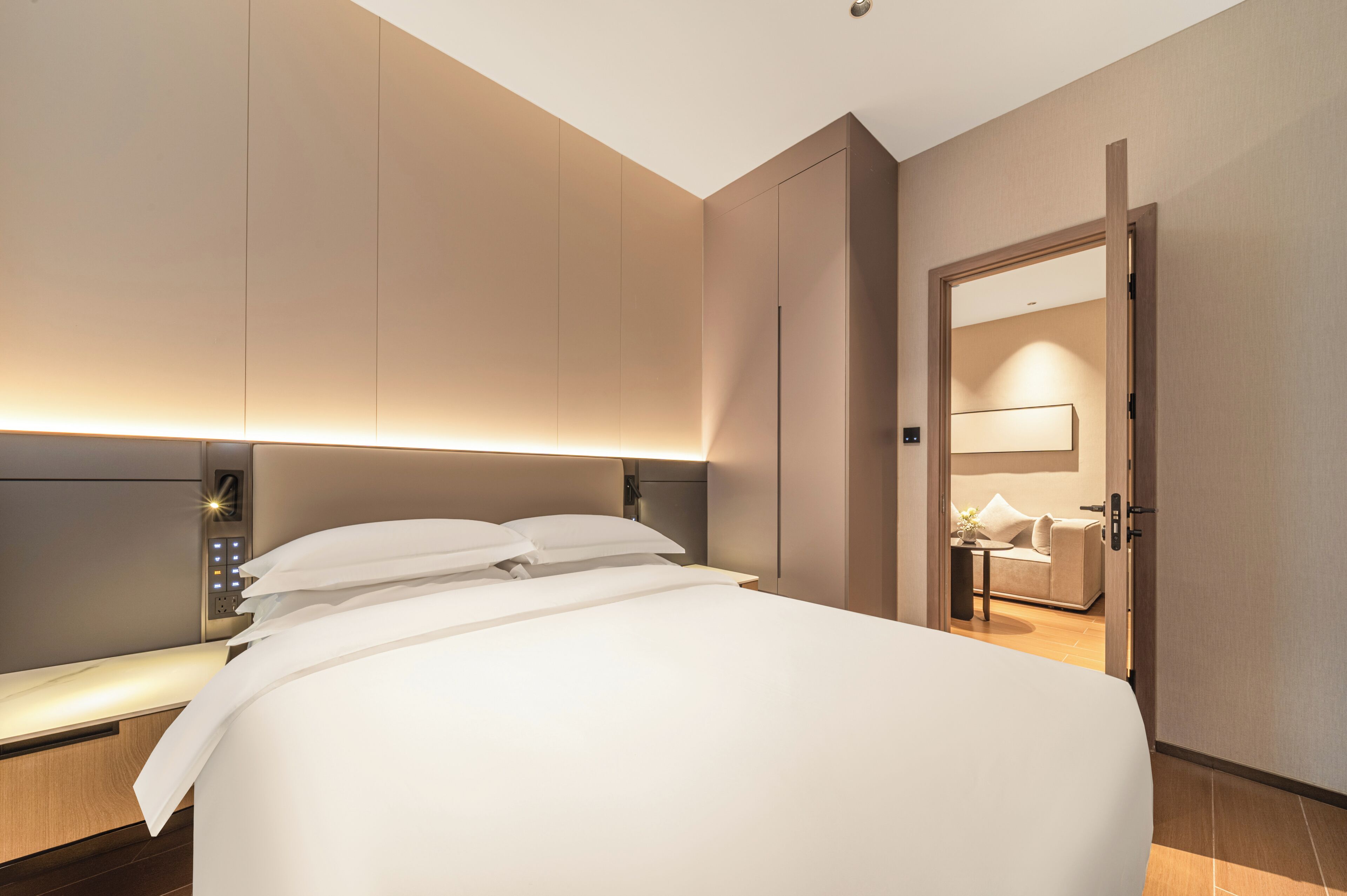 A modern hotel room with a large bed, bedside tables, a nightstand, a wall-mounted lamp, and a door leading to a bathroom.