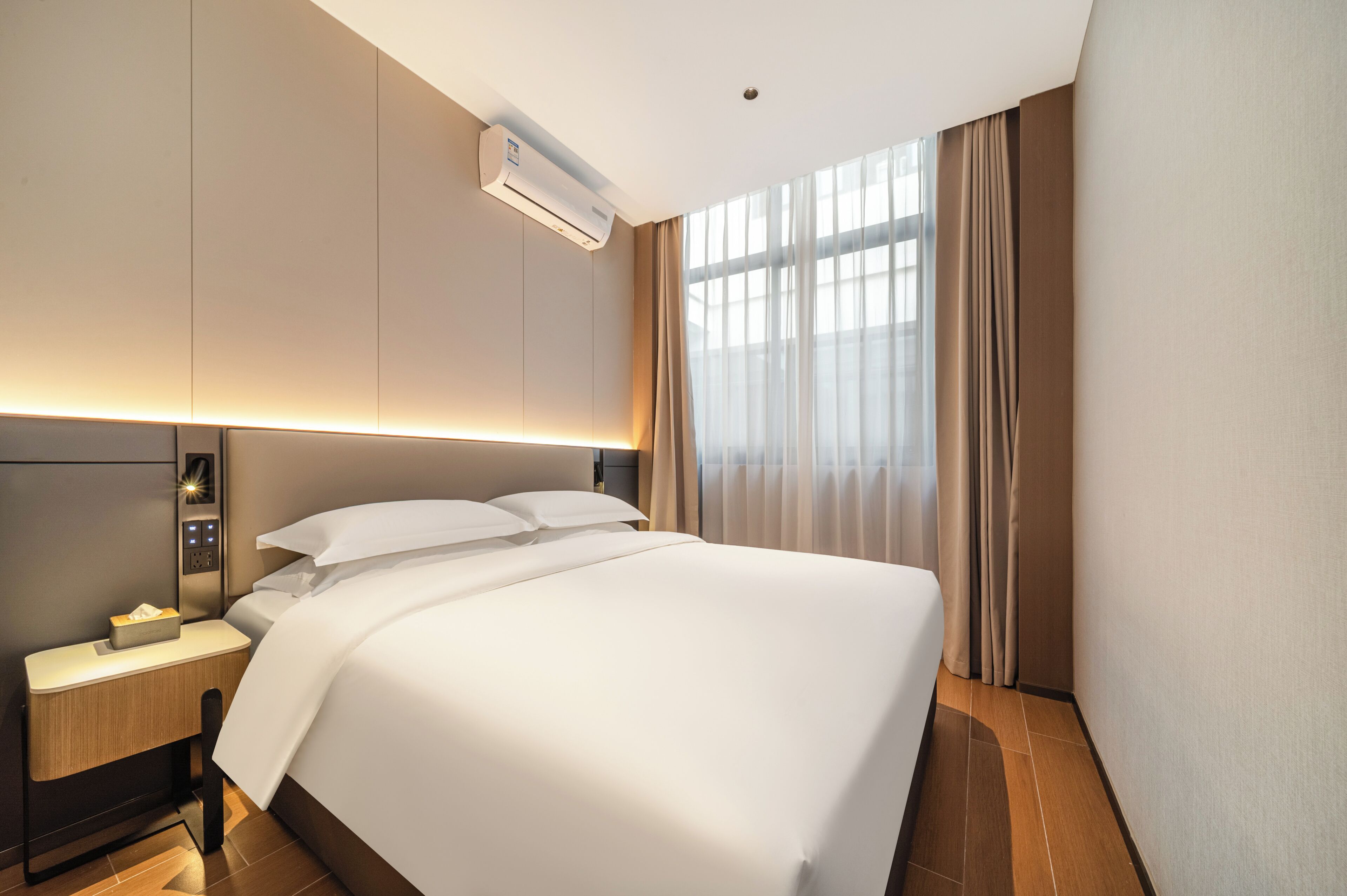 A hotel room with a large bed, a bedside table with a lamp, an air conditioning unit, and a window with curtains.