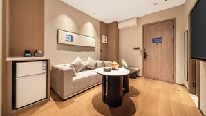 A modern hotel room with a sofa, a small table with a bowl of fruit, a mini-fridge, a flat-screen TV, and a door with a room number sign.