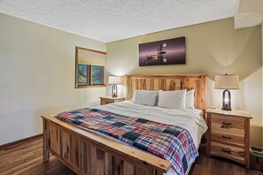 A bedroom with a wooden bed, a nightstand with a lamp, a wall-mounted TV, and a painting. - Lakefront 4BR/3.5BA retreat with beautiful views (Oakland)