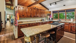 A kitchen with wooden cabinets, granite countertops, a central island with stools, and a view through large windows. - Lakefront 4BR/3.5BA retreat with beautiful views (Oakland)