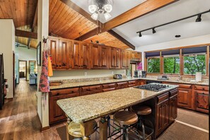 A kitchen with wooden cabinets, granite countertops, a central island with stools, and a view through large windows. - Lakefront 4BR/3.5BA retreat with beautiful views (Oakland)