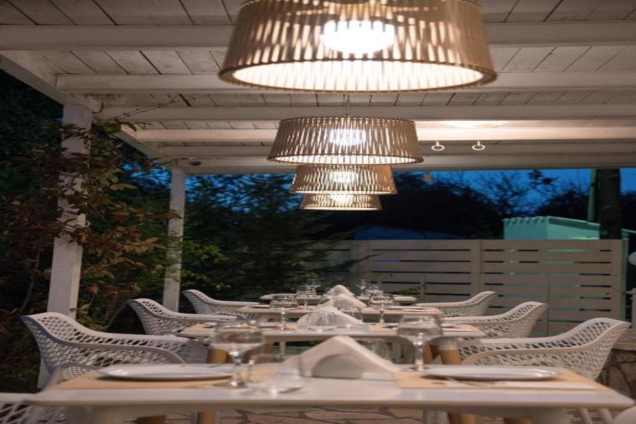Ein Essbereich im Freien mit weißen Korbstühlen und Tischen, unter einer Pergola mit hängenden Lampen.