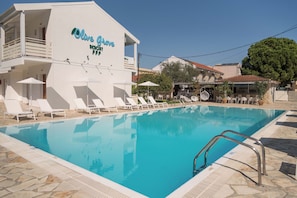 Ein Schwimmbecken mit Liegestühlen und Sonnenschirmen, ein Gebäude mit der Aufschrift 'Olive Grove Resort' und ein klarer, blauer Himmel.