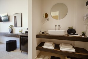 A modern bathroom with a large white sink, a round mirror, and a hairdryer on the right. There are towels on the shelves and a black trash bin to the left. - Eleonas Bohotel & Annex Apartments (Corfu)