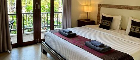A hotel room with a large bed, a nightstand with a lamp, a ceiling fan, and a view of greenery through the sliding glass doors.