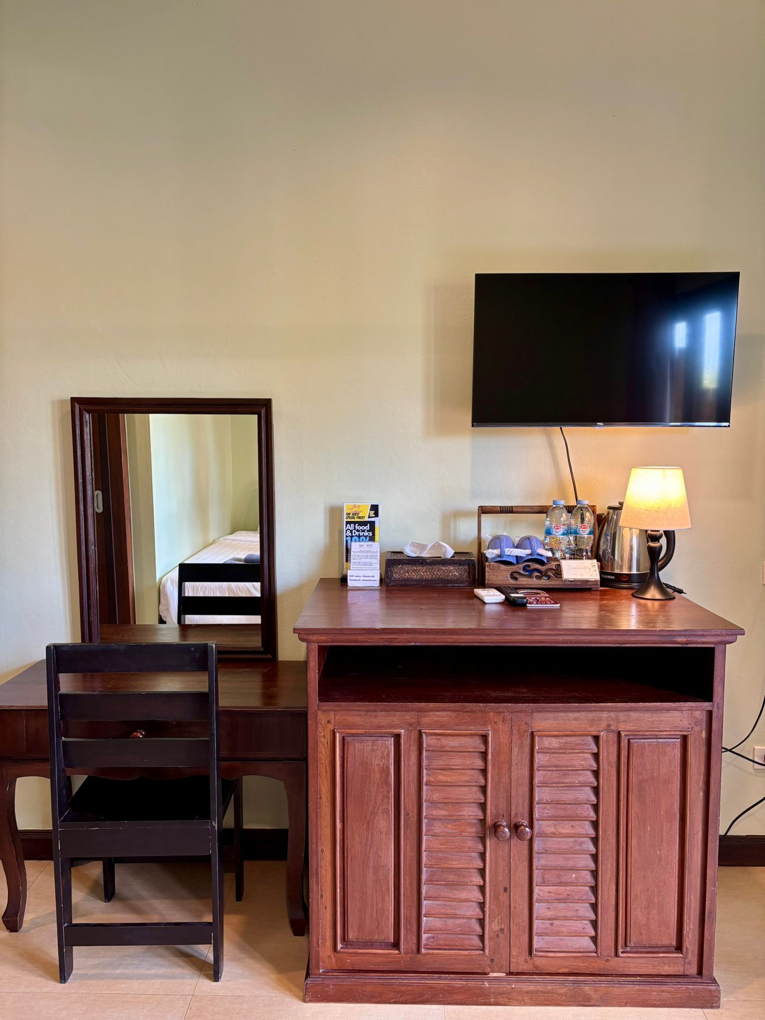 Habitación de hotel con escritorio de madera, una silla, televisor montado en la pared y una mesita de noche con lámpara.