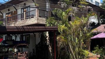 A two-story building with a balcony, a parked tuk-tuk, and a garden area with various plants.