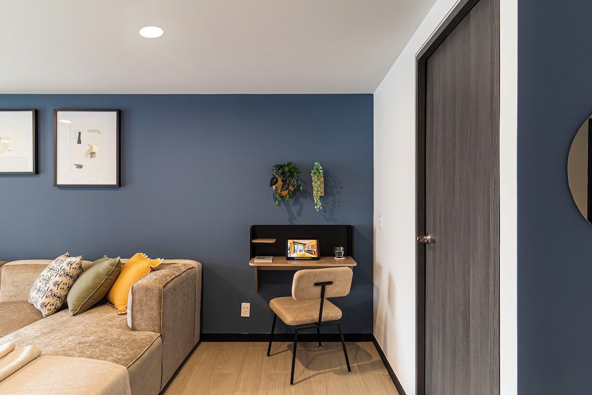 A modern room with a dark blue wall, a sofa with cushions, a small desk with a laptop, and a door.