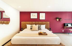Un dormitorio moderno con una cama grande, cabecera, dos cuadros enmarcados, una mesita de noche con un ordenador portátil y una planta pequeña.