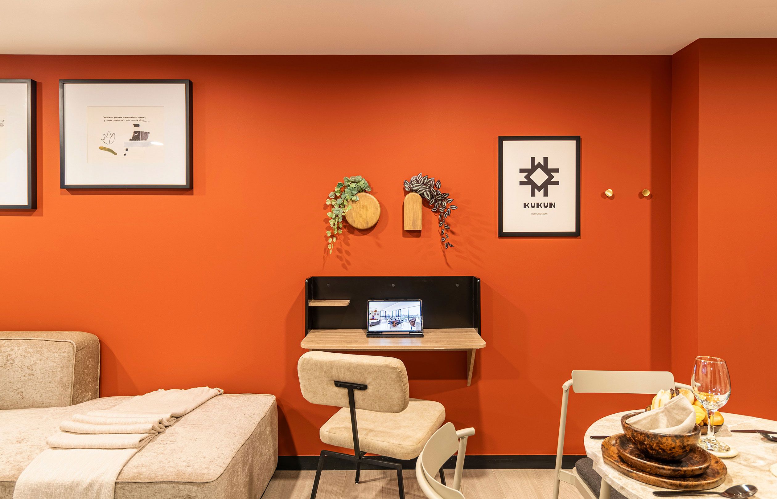 Habitación de hotel con una cama, un escritorio con una computadora portátil, una silla, una mesita con un tazón y un vaso, y cuadros en la pared.
