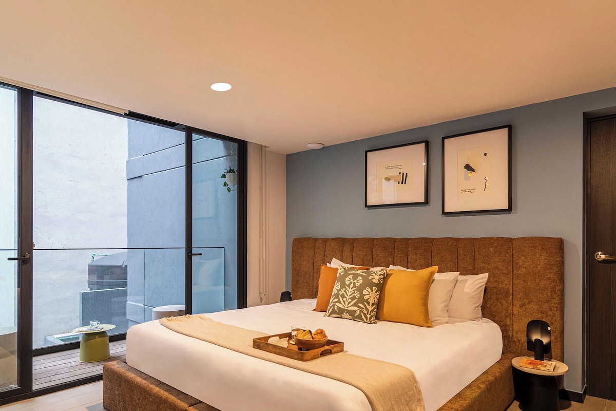 Un dormitorio moderno con una cama grande, una mesita de noche, una pequeña mesa redonda y un ventanal corredero que da a un balcón.
