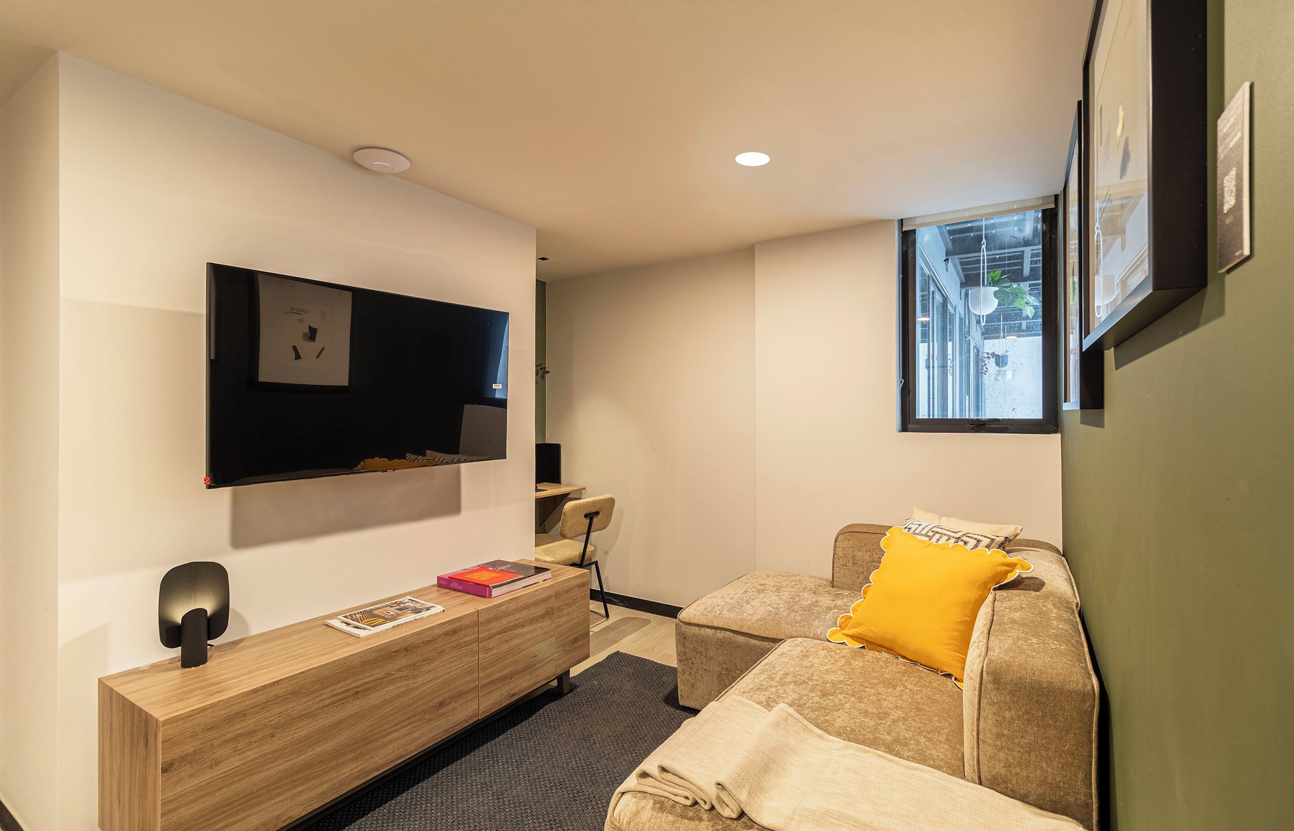 Una sala moderna con un televisor de pantalla plana montado en la pared, una consola de madera con libros, un sofá marrón con un cojín amarillo y una ventana que da a la calle.