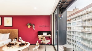 A modern interior with a red accent wall, a dining area with a table set for two, a workspace with a laptop, and a balcony with a view of the city. - Casa Sócrates By Kukun (Mexico City)