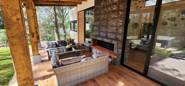 An outdoor living area with a glass enclosure, wicker furniture, and a stone fireplace. - Cima Boreal Chalet (Tapalpa)