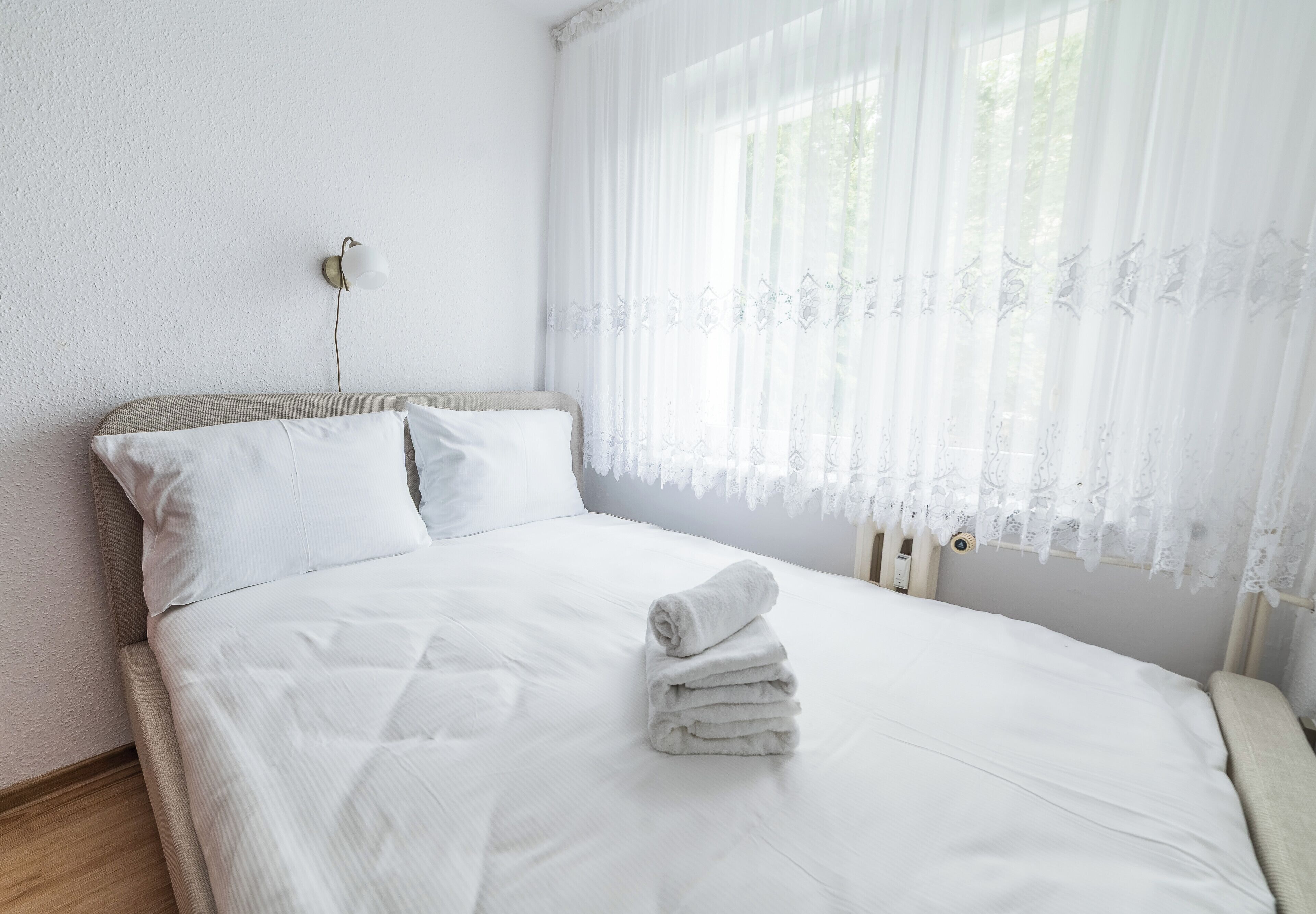 Una cama bien hecha con sábanas blancas y una pila de toallas, un aplique en la pared y cortinas de encaje con estampado floral.