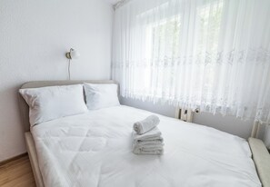 A neatly made bed with white linens and a stack of towels, a wall-mounted light fixture, and sheer curtains with a floral pattern. - Pod Lipami Cozy Apartment (Poznań)