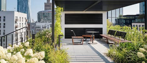 A rooftop terrace with a seating area, surrounded by greenery and overlooking a cityscape.