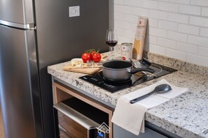 Eine Küche mit einem Edelstahl-Kühlschrank, einer schwarzen Induktionskochplatte, einem Schneidebrett mit Käse und Tomaten, einem Weinglas und einer Bierdose.