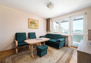 Ein Wohnzimmer mit einer Couch, Sesseln, einem kleinen Tisch, einem Gemälde und Blick auf die Stadt durch große Fenster.