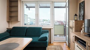 A living room with a green sofa, a wooden cabinet, a television, and a view of the city through large windows. - Solec Powiśle Standard Apartment (Warszawa)