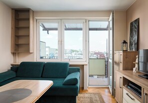 Ein Wohnzimmer mit einem grünen Sofa, einer Holzkommode, einem Fernseher und Blick auf die Stadt durch große Fenster.