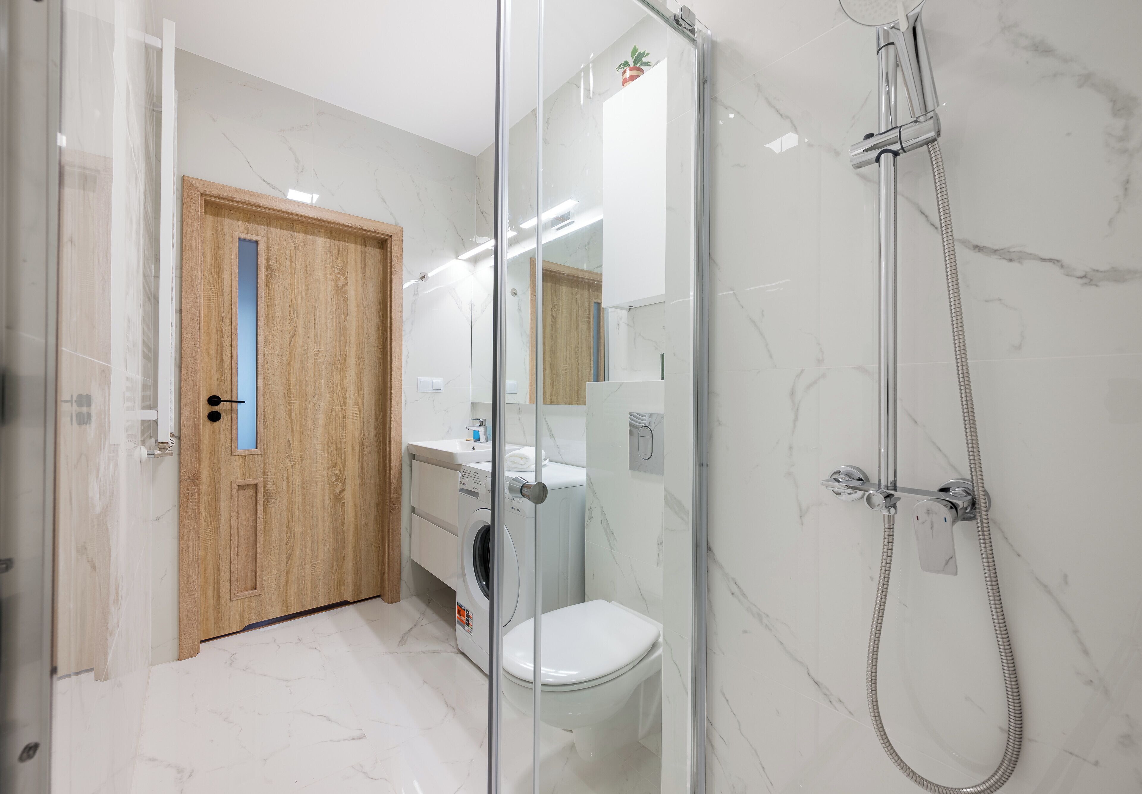 Un baño moderno con cabina de ducha de vidrio, puerta de madera, inodoro blanco y suelo de baldosas de mármol.